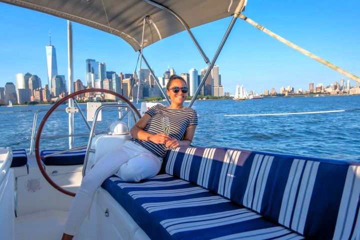 woman sitting on a private boat charter in NYC. Seating has blue stripes. Behind her is the steering wheel and Manhattan skyline.